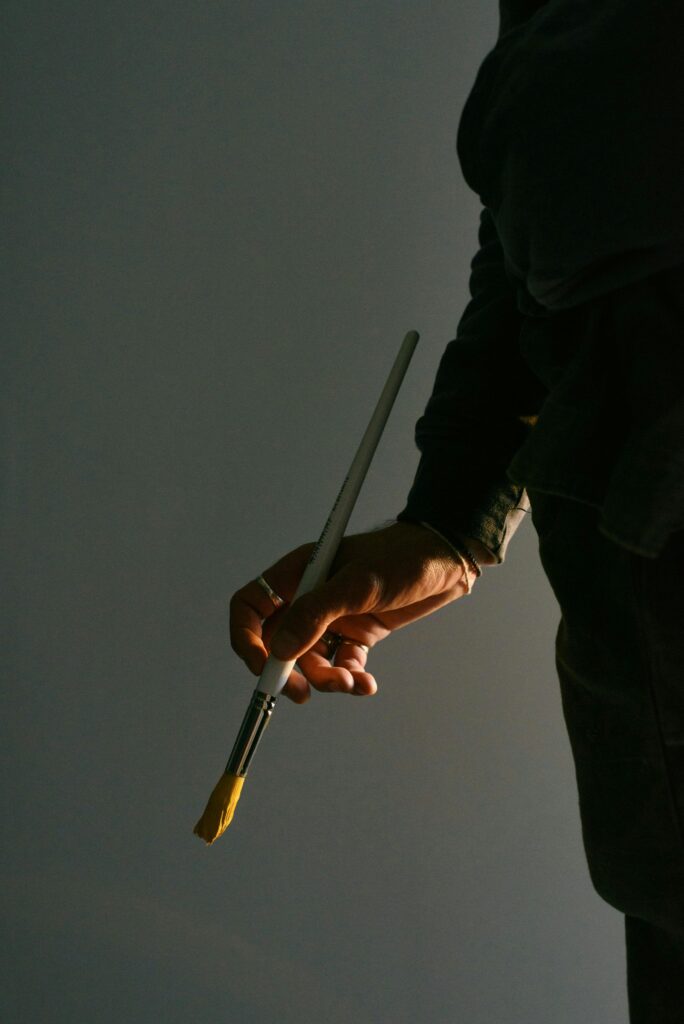A close-up of an artist's hand holding a yellow paintbrush against a gray background.